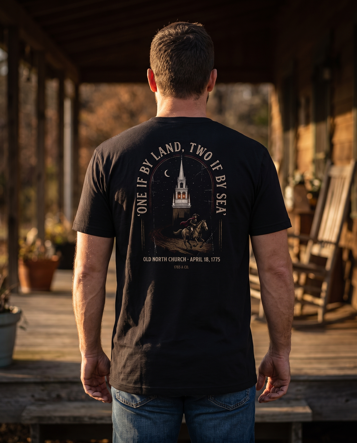 Man seen from behind wearing a tee with the Old North Church steeple, two lit lanterns and a galloping rider on the back