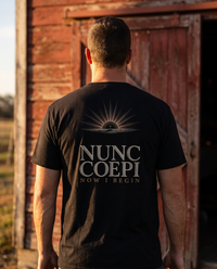 Man seen from behind in a tee whose back print shows a gold cross in a sunburst over NUNC COEPI and NOW I BEGIN &mdash; view 1