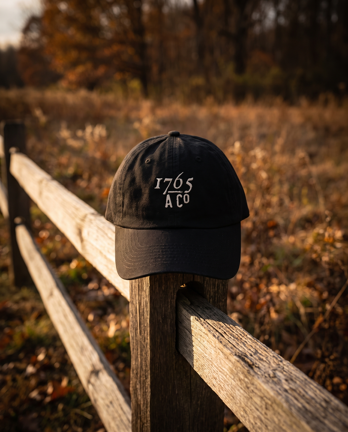 1765 A Co. dad cap in black with the embroidered mark, resting on a split-rail fence at golden hour
