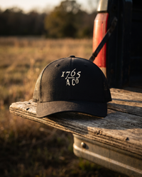 1765 A Co. trucker cap in black with the embroidered mark and mesh back, on a pickup tailgate at golden hour &mdash; view 1